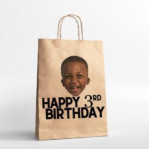 K&ouml;nnte beinhalten: Eine braune Papiert&uuml;te mit einem Foto eines jungen Jungen auf der Vorderseite. Die Tasche hat den Text "Happy 3rd Birthday" in Schwarz aufgedruckt.