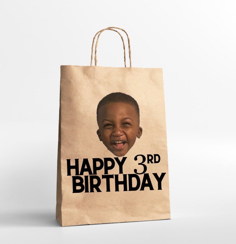 K&ouml;nnte beinhalten: Eine braune Papiert&uuml;te mit einem Foto eines jungen Jungen auf der Vorderseite. Die Tasche hat den Text "Happy 3rd Birthday" in Schwarz aufgedruckt.