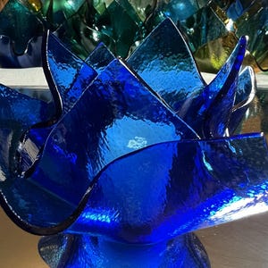 May include: A vibrant, deep blue glass bowl with a unique, petal-like design. The bowl's edges are sharply angled, creating a dynamic, sculptural form. Other bowls in shades of green and blue are visible in the background.