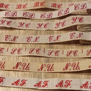 Könnte beinhalten: Mehrere Spulen und Längen von Vintage-Bändern mit roten, gestickten Initialen. Die Bänder sind cremefarben und cremeweiß, mit verschiedenen Initialen wie "B.C.", "C.D." und "A.F.". Geeignet für Näh- oder Bastelprojekte.