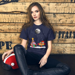 Puede incluir: Una camiseta azul marino con un árbol de Navidad y un taco con un gorro de Papá Noel. El árbol está decorado con adornos coloridos y el taco sonríe. La luna está en el fondo.