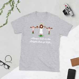 Könnte beinhalten: Ein heidefarbenes T-Shirt mit einem Cartoon-Bild von drei Personen auf Kreuzen. Der Text "Always look on the bright side of life..." ist unter dem Bild gedruckt.