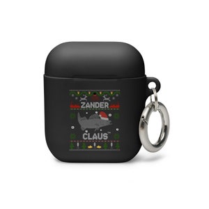 Könnte beinhalten: Schwarze Airpods-Hülle mit einem Weihnachtsdesign, das einen Fisch mit einem Weihnachtsmann-Hut und dem Text "Zander Claus" zeigt. Die Hülle hat einen silbernen Metall-Schlüsselanhänger.