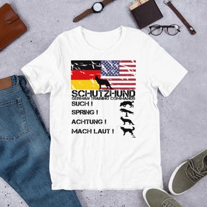 Könnte beinhalten: Weißes T-Shirt mit einem abgenutzten Design der amerikanischen und deutschen Flagge. Unter den Flaggen steht der Text "Schutzhund German Training Commands", gefolgt von den Befehlen "Such!", "Spring!", "Achtung!" und "Mach Laut!" mit entsprechenden Hundesilhouetten.