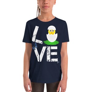 K&ouml;nnte beinhalten: Ein marineblaues T-Shirt f&uuml;r Jugendliche mit einem wei&szlig;en, abgenutzten Aufdruck des Wortes "LOVE" mit einem K&uuml;ken, das aus einem Ei im "O" schl&uuml;pft. Ein blauer Schmetterling befindet sich in der oberen linken Ecke des Aufdrucks.
