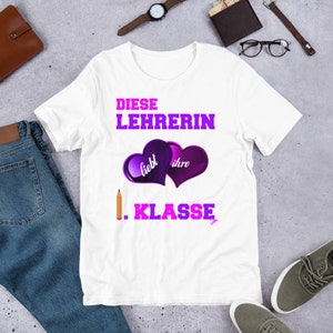 Puede incluir: Camiseta blanca con un gráfico rosa y morado que dice "Diese Lehrerin liebt ihre Klasse" con un lápiz y dos corazones.