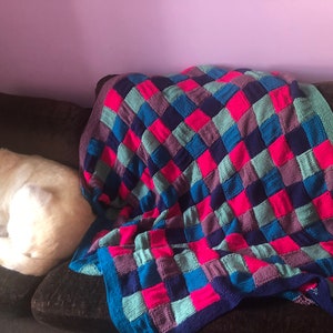 Puede incluir: Un perro blanco está durmiendo en un sofá marrón con una manta de punto de colores drapeada sobre el respaldo del sofá. La manta está hecha de cuadrados en tonos de rosa, azul, verde y morado.