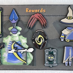 Może przedstawiać: Zestaw dziewięciu emaliowanych pinów przedstawiających różne postacie i przedmioty z gry wideo. Piny są wyświetlane na szarym tle z napisem "Rewards" wydrukowanym na pomarańczowo. Piny obejmują łuk i strzałę, niebiesko-fioletową istotę, czerwoną szponę, czarno-żółte oko, niebiesko-biały kapelusz czarodzieja, niebiesko-białą szatę, czarno-zieloną tarczę, niebiesko-biały płaszcz i czarno-biały młotek.