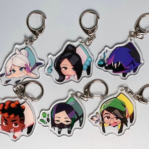 May include: Set of six colorful character keychains with different designs. Each keychain features a cartoon character with a unique expression and pose. The keychains are made of clear acrylic and have silver metal key rings.