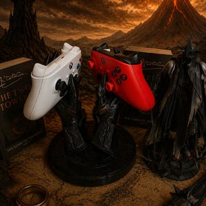 May include: A white and a red Xbox controller are displayed on black stands, set against a backdrop of a fantasy landscape with a volcano and a dark tower. The scene includes a book, a ring, and a figurine.