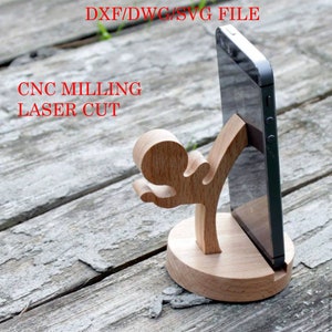 May include: A wooden phone stand shaped like a person kicking a phone. The stand is made of light brown wood and has a round base. The phone is black and has a white screen. The text "DXF/DWG/SVG FILE CNC MILLING LASER CUT" is written in red letters at the top of the image.