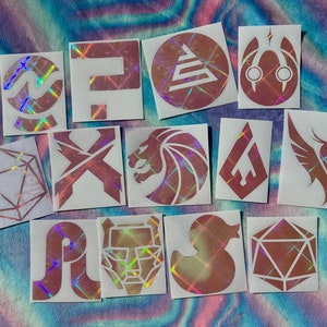 Puede incluir: Un conjunto de 12 pegatinas holográficas iridiscentes con varios símbolos y diseños. Las pegatinas están sobre un fondo blanco y tienen un brillo iridiscente rosa, azul y morado.