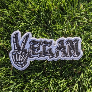 May include: Embroidered patch with the word "VEGAN" in black letters with a skeleton hand making a peace sign. The patch has a white border and is set against a background of green grass.