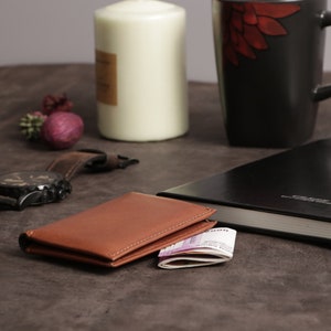 May include: A brown leather wallet with a stack of bills inside, a black leather journal with a blue pen, a black wristwatch with a brown leather band, and a white candle on a brown surface.