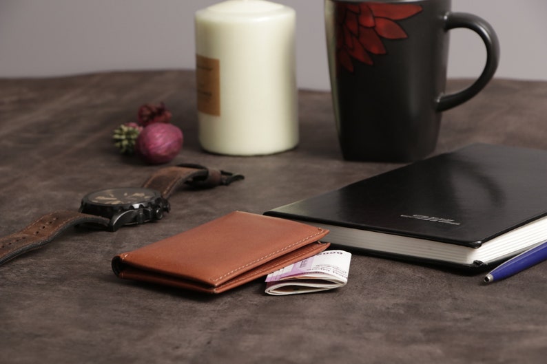 May include: A brown leather wallet with a stack of bills inside, a black leather journal with a blue pen, a black wristwatch with a brown leather band, and a white candle on a brown surface.