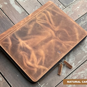May include: A brown leather zippered portfolio with a subtle distressed look. The portfolio has a stitched edge and a zipper pull with a metal tag that says "ALASKA". The text "NATURAL CAMEL" is visible at the bottom of the image.