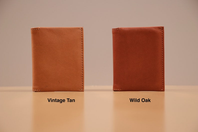 May include: Two leather wallets in different colors. The wallet on the left is a light brown color labeled "Vintage Tan". The wallet on the right is a reddish brown color labeled "Wild Oak".
