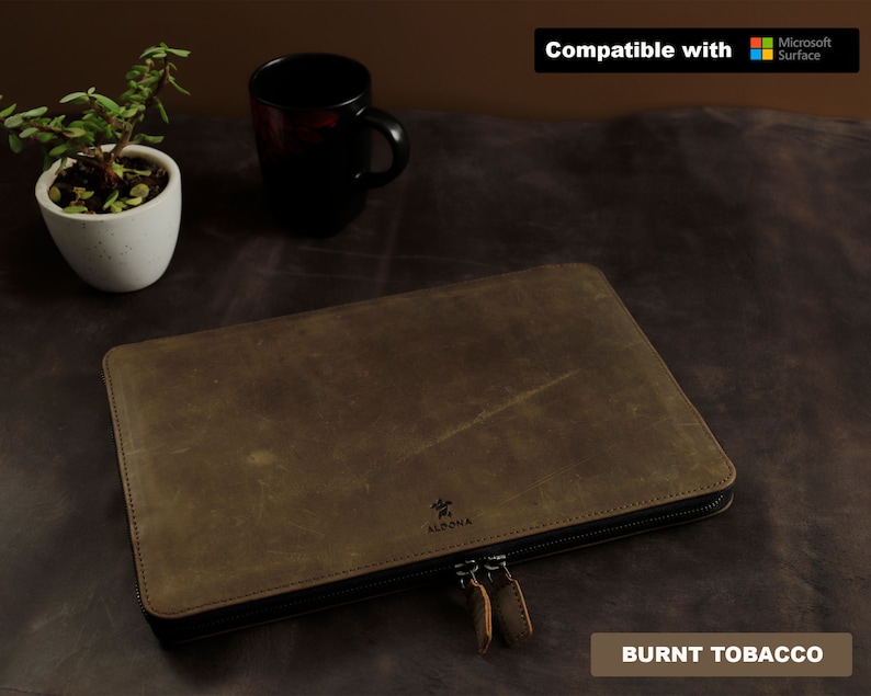 May include: A brown leather laptop sleeve with a zipper closure. The sleeve has a logo that says "Aldona" and the text "Burnt Tobacco" is visible at the bottom of the image. The sleeve is compatible with Microsoft Surface.