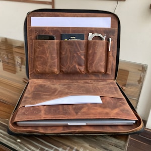 May include: Open brown leather laptop case with multiple pockets and a zipper closure. The case is open and a laptop is inside. There are pockets for a phone, passport, and other items.
