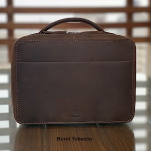 May include: A brown leather zipped case with a small logo that says "Aurora". The case is on a wooden surface and the text "Burnt Tobacco" is below the case.