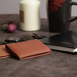 May include: Two brown leather wallets, one on top of the other, on a brown textured surface. A black leather journal with a blue pen is in the background.