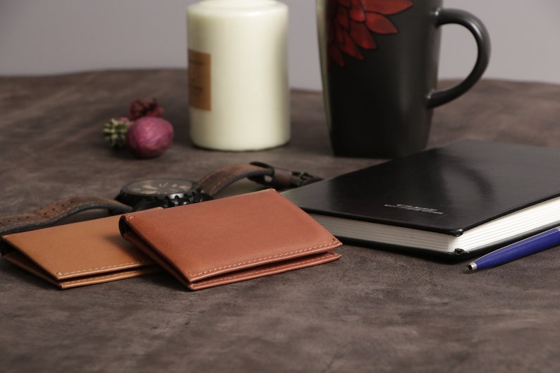 May include: Two brown leather wallets, one on top of the other, on a brown textured surface. A black leather journal with a blue pen is in the background.