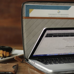 May include: A gray laptop case with a zipper closure. The case is open and a laptop is inside. The laptop screen shows a website with the text "iPhone X from $10.20/mo. or $249". The case has a brown leather zipper pull and a brown leather pocket on the front. The case is sitting on a wooden table with a donut on a white plate.