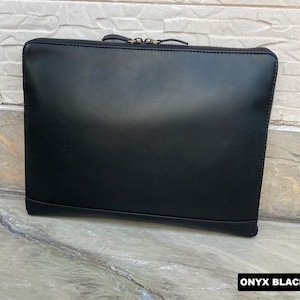 May include: A black leather laptop sleeve with a zipper closure. The sleeve is on a light gray textured surface. "ONYX BLACK" is written in white text on the surface.