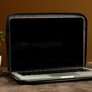 May include: A black laptop computer in a black leather case with a zipper. The laptop is open and the screen is black. The laptop is sitting on a brown surface.