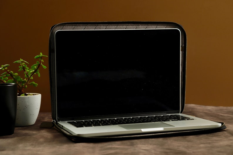 May include: A black laptop computer in a black leather case with a zipper. The laptop is open and the screen is black. The laptop is sitting on a brown surface.