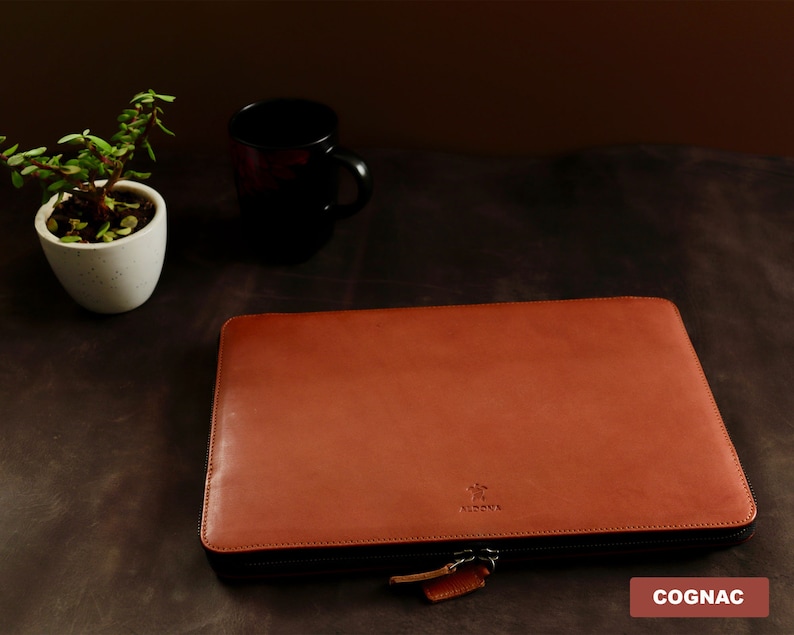 May include: A cognac-colored leather laptop sleeve with a zipper closure. The sleeve has a small logo that says "Artova" on the front.
