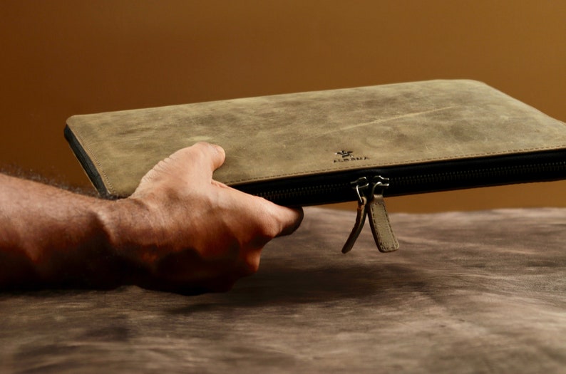 May include: A brown leather laptop sleeve with a zipper closure and a small logo on the front. The sleeve is being held by a hand.