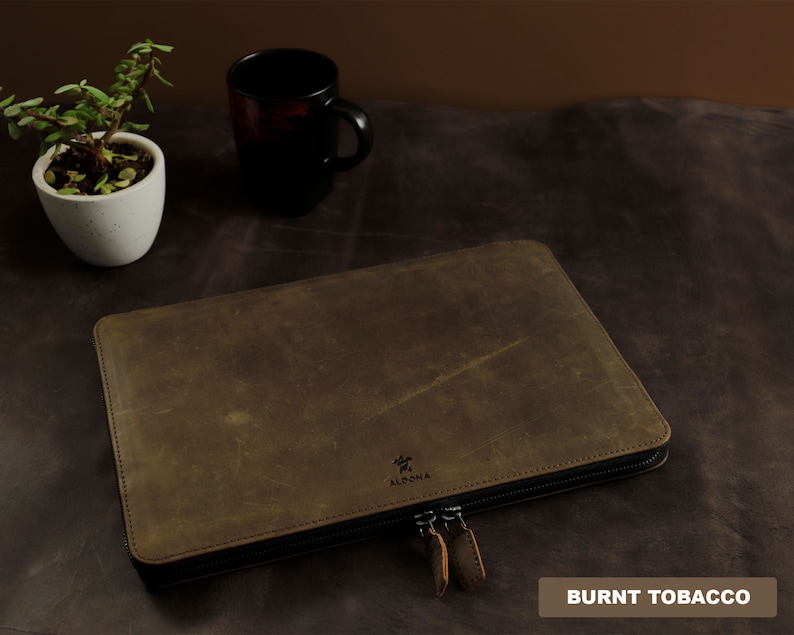 Puede incluir: Una funda de cuero marr&oacute;n con cremallera con la palabra "ALDONA" grabada en la parte delantera. La funda est&aacute; etiquetada como "BURNT TOBACCO".