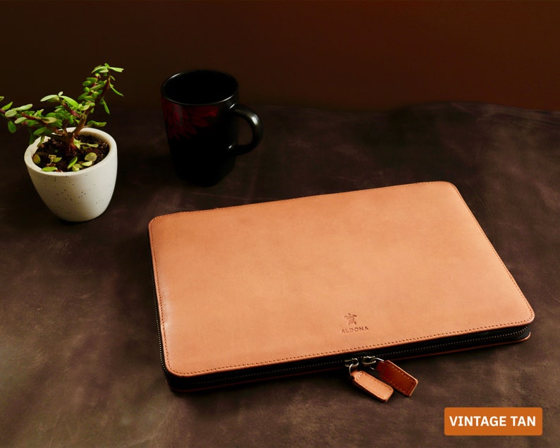 May include: A vintage tan leather laptop sleeve with a zipper closure and the word "ALDONA" embossed on the front.