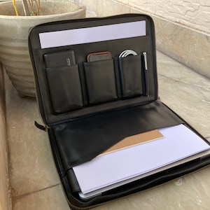 Può includere: Custodia per laptop in pelle nera con più tasche e chiusura a zip. La custodia è aperta e contiene un laptop, un blocco note, un telefono e un caricabatterie.