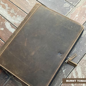 May include: A brown leather zippered portfolio with a stitched edge and a small logo on the front. The portfolio is on a wooden surface. BURNT TOBACCO