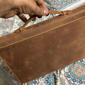 Può includere: Una valigetta in pelle marrone con una maniglia superiore. La valigetta ha un aspetto usurato e invecchiato.