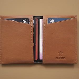May include: A brown leather wallet with a logo that says "ALDONA" and a turtle design. The wallet is open and has several cards and cash inside.