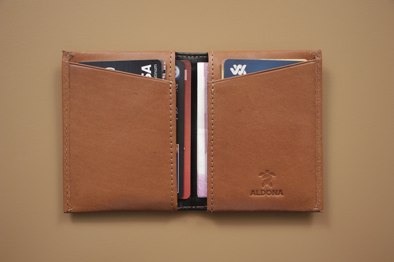 May include: A brown leather wallet with a logo that says "ALDONA" and a turtle design. The wallet is open and has several cards and cash inside.