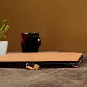 May include: A tan leather laptop sleeve with a zipper closure. The sleeve is lying on a brown fabric surface.