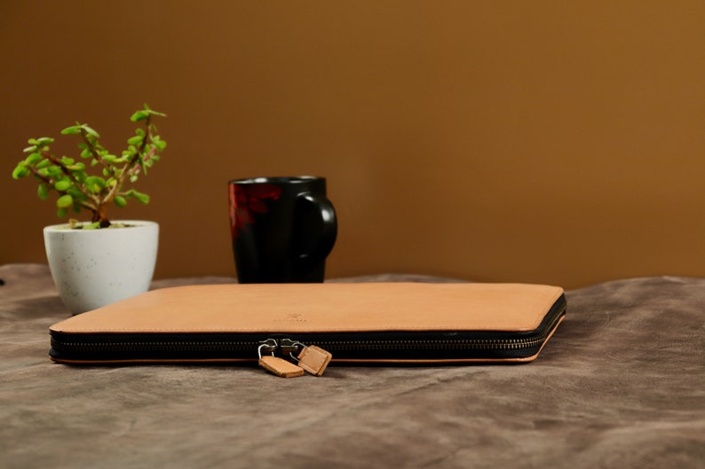 May include: A tan leather laptop sleeve with a zipper closure. The sleeve is lying on a brown fabric surface.