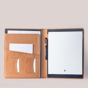 Könnte beinhalten: Geöffnetes Portfolio aus hellbraunem Leder mit schwarzem Rand, das weißes Papier und einen schwarzen Stift enthält. Das Portfolio hat Taschen für Karten und Dokumente. Ein kleines schwarzes Logo ist auf dem weißen Papier sichtbar.