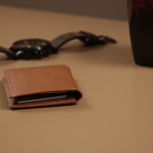 May include: A brown leather wallet with a folded design. The wallet is closed and sitting on a light brown surface.