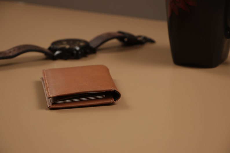 May include: A brown leather wallet with a folded design. The wallet is closed and sitting on a light brown surface.