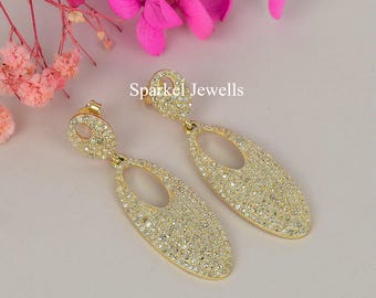 May include: A pair of gold-toned dangle earrings. Each earring features an oval shape with a central cutout, encrusted with small, sparkling stones. The earrings are displayed on a light pink surface, with pink flowers in the background. The text "Sparkel Jewells" is visible.