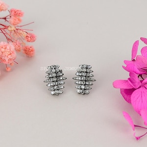 Op de afbeelding: Een paar zilverkleurige oorstekers. Elke oorbel heeft een gestapeld, langwerpig ontwerp met meerdere rijen kleine, heldere edelstenen. De oorbellen staan tegen een zachtroze achtergrond, met roze bloemen in de hoeken.
