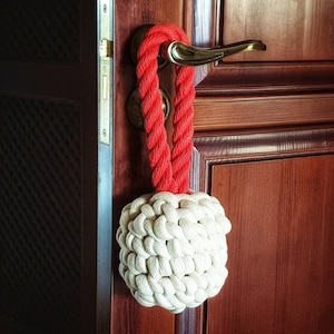 May include: A red rope doorstop with a white, crocheted ball. The doorstop is hanging on a wooden door.