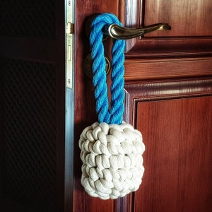 Pode incluir: Um batente de porta de corda azul com uma bola de crochê branca. O batente de porta está pendurado em uma porta de madeira.