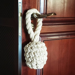 Op de afbeelding: Een witte touwdeurstopper met een gevlochten ontwerp. De deurstopper hangt aan een deurknop aan een houten deur.