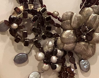 May include: A multi-strand necklace with a silver flower charm, dark red and white beads, and a silver circle charm with dark red stones.